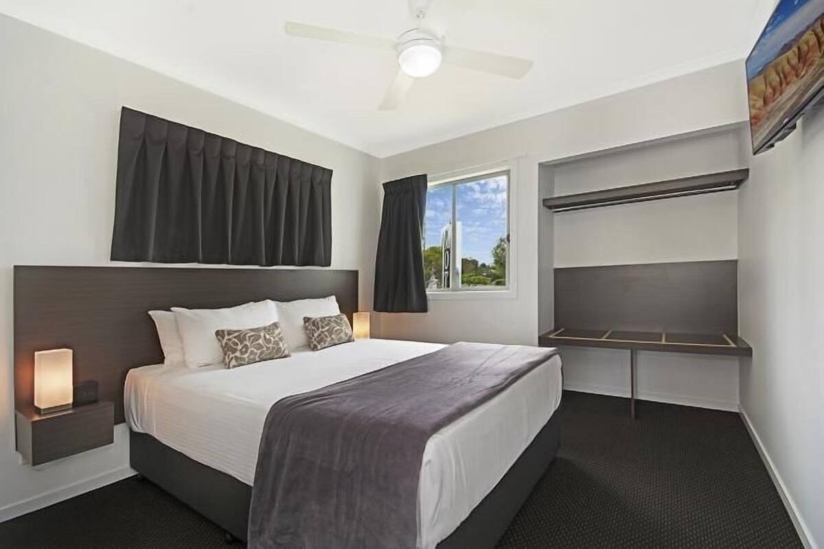 A hotel room with a bed, bedside lamp, ceiling fan, window with curtains, and a built-in shelf.