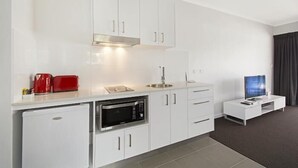 A modern kitchen with white cabinets, a built-in oven, a microwave, a coffee maker, and a TV on a white stand. - Cooroy Luxury Motel Apartments Noosa (Cooroy)