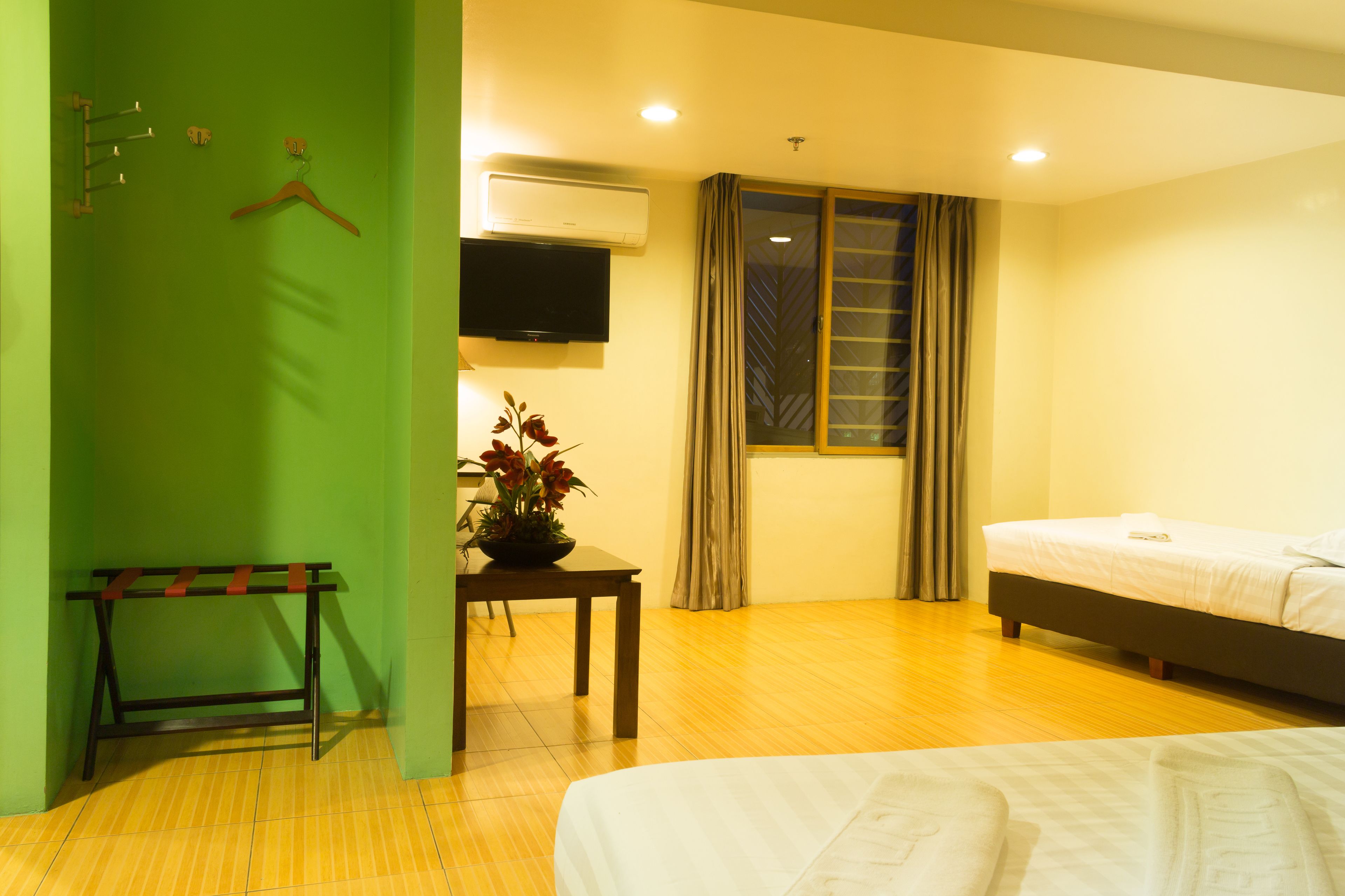 Una habitación de hotel con cama, una mesa con un jarrón de flores, perchero, televisor y ventana con cortinas.