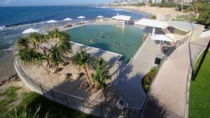 Un espace piscine offrant une vue sur la plage et l’océan, entouré de palmiers et de parasols.