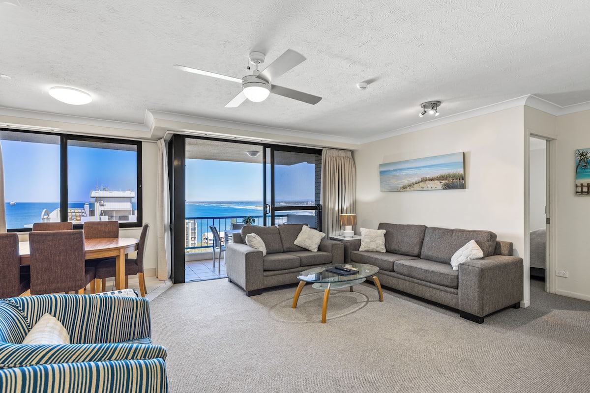 Een moderne woonkamer met een bank, fauteuils en een salontafel. Er is een balkon met uitzicht op zee.