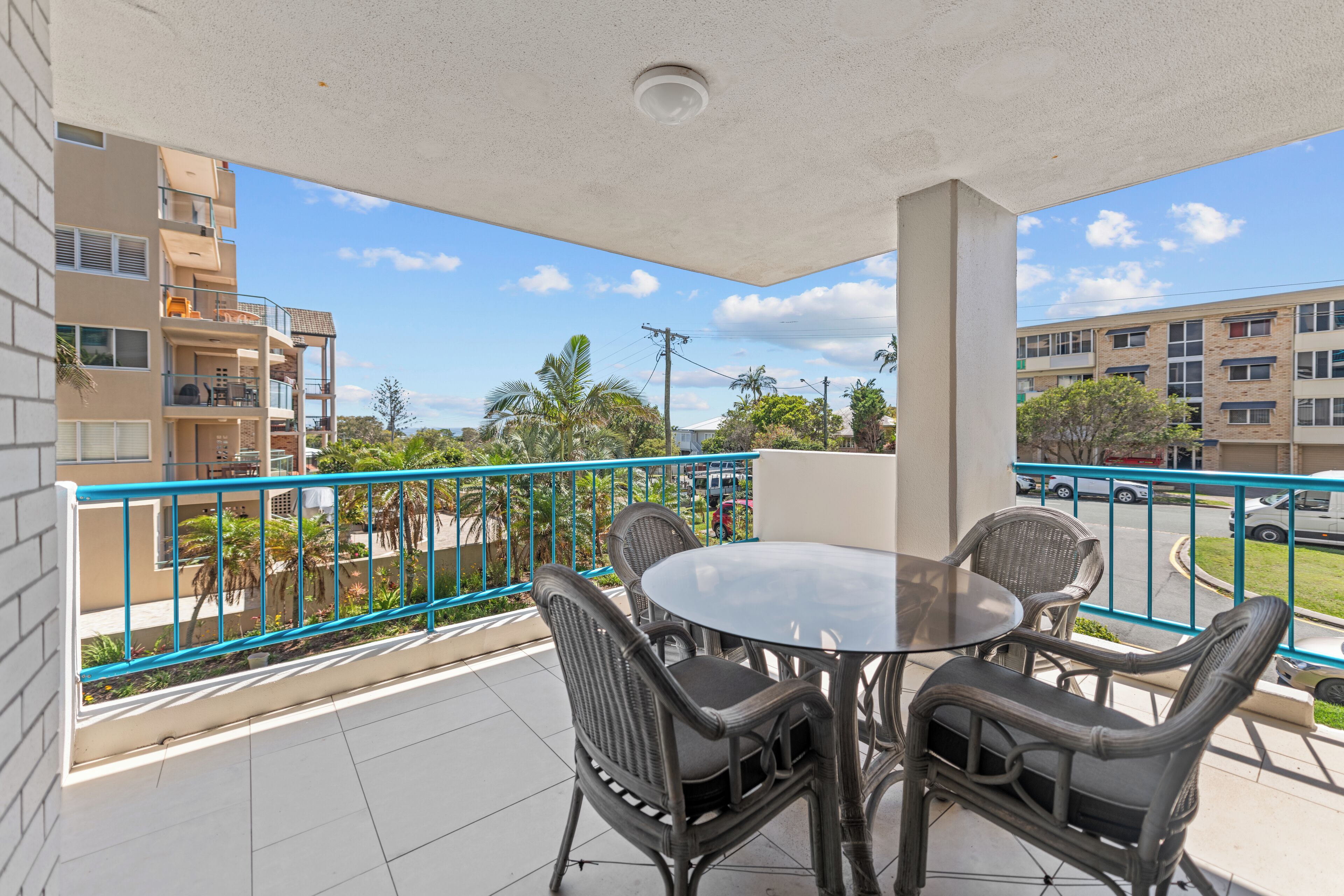 A balcony with a round table and four chairs, overlooking a street with buildings and trees.
