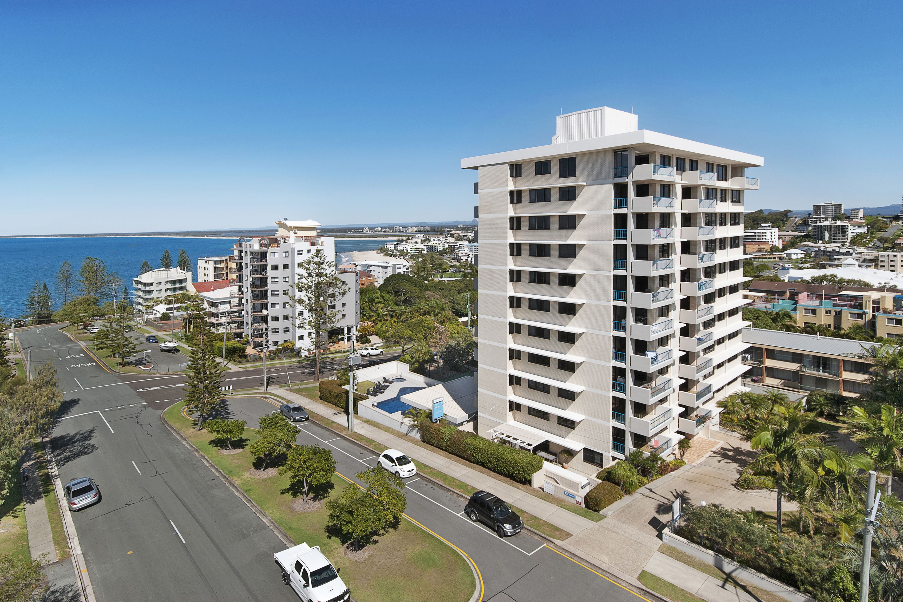 A high-rise hotel with multiple balconies, a parking lot, and a view of the sea.