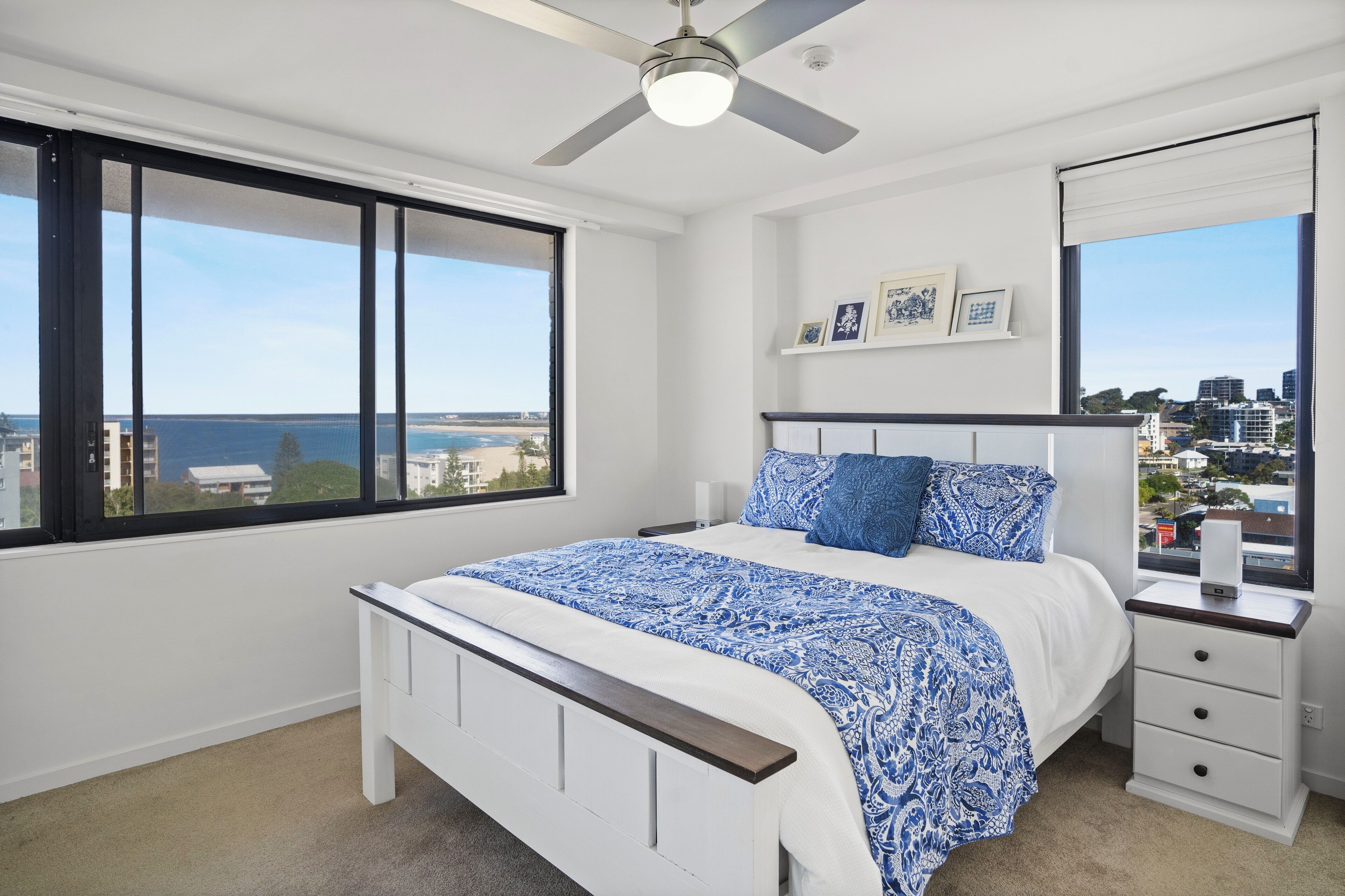 A bedroom with a bed, a nightstand, a ceiling fan, a large window with a view of the city, and a wall-mounted shelf with pictures.