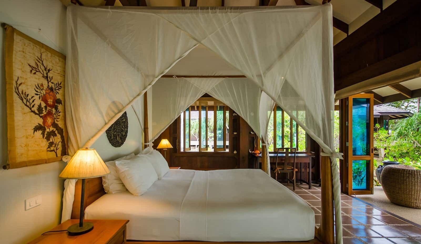 A bedroom with a canopy bed, a nightstand with a lamp, a chair, and a view of the garden through the window.