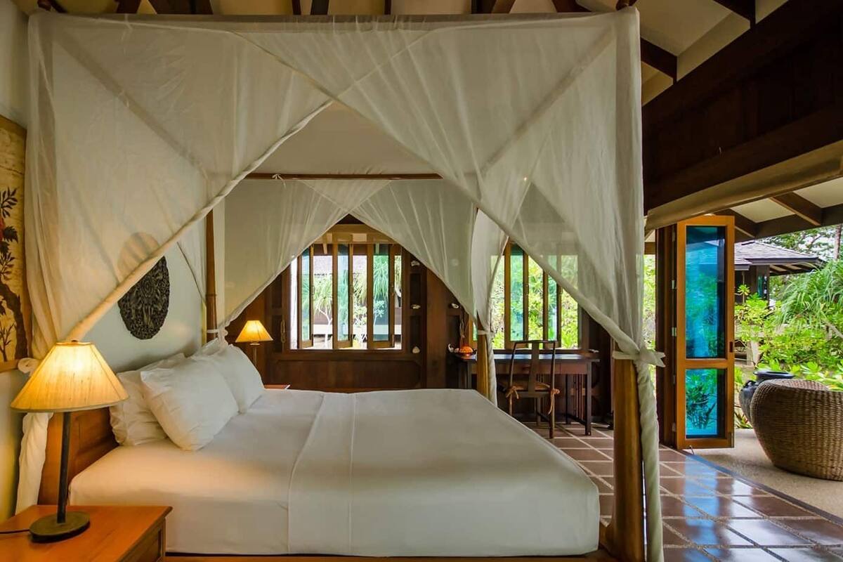Une chambre à coucher avec un lit à baldaquin, une table de chevet avec une lampe, un fauteuil et une vue sur le jardin par la fenêtre.