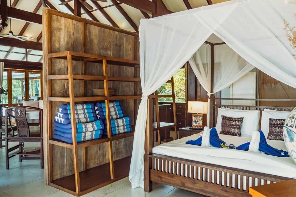 Une chambre avec un lit, une étagère en bois où sont pliées des couvertures et un lit à baldaquin.