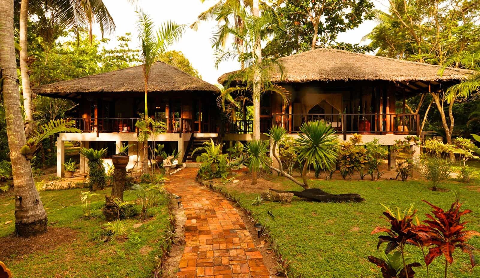 A tropical resort with two thatched-roof cabins, a paved pathway, and lush greenery.