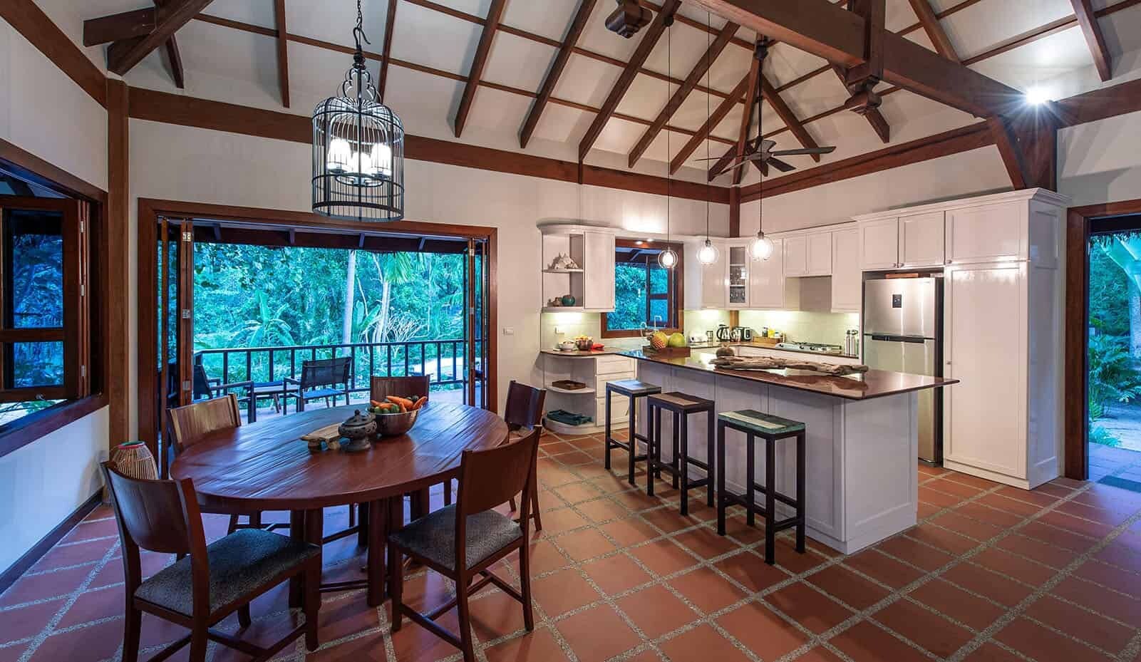 A spacious kitchen with a large island, wooden dining table, and chairs, with a view of greenery outside.