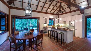 A spacious kitchen with a large island, wooden dining table, and chairs, with a view of greenery outside.