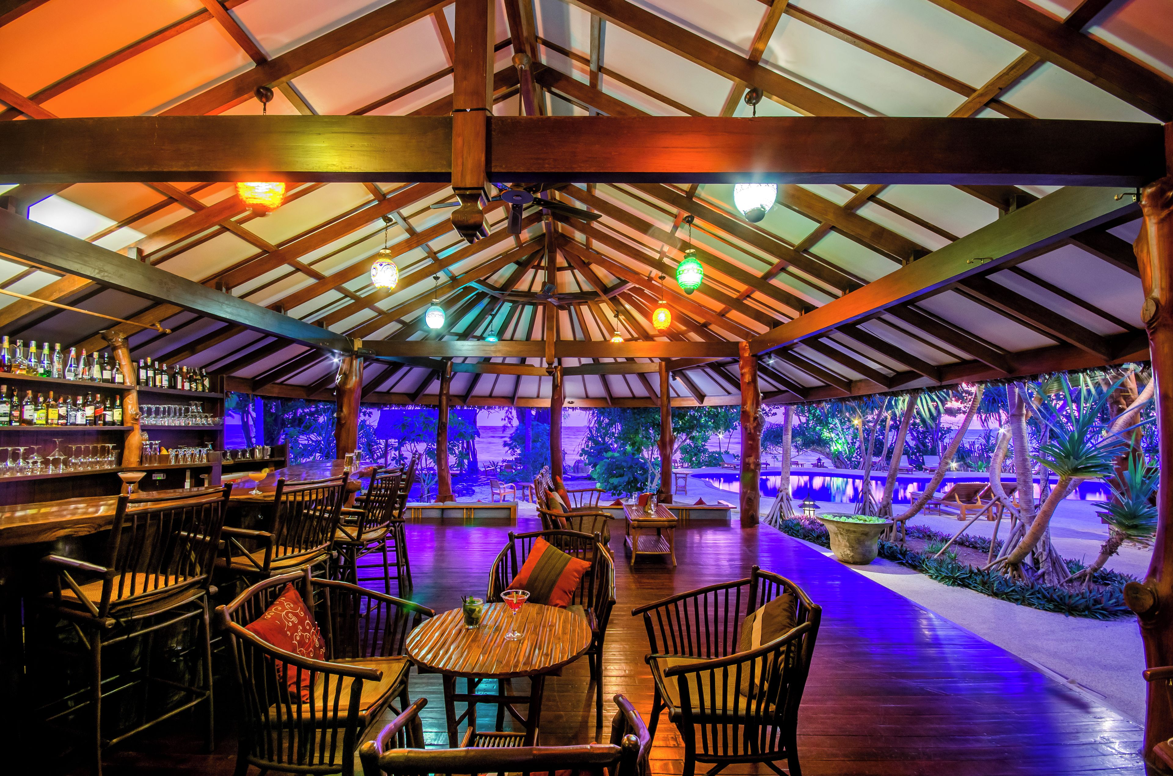 Un'area bar all'aperto con mobili in legno, un bancone bar pieno di bottiglie e vista sugli alberi.