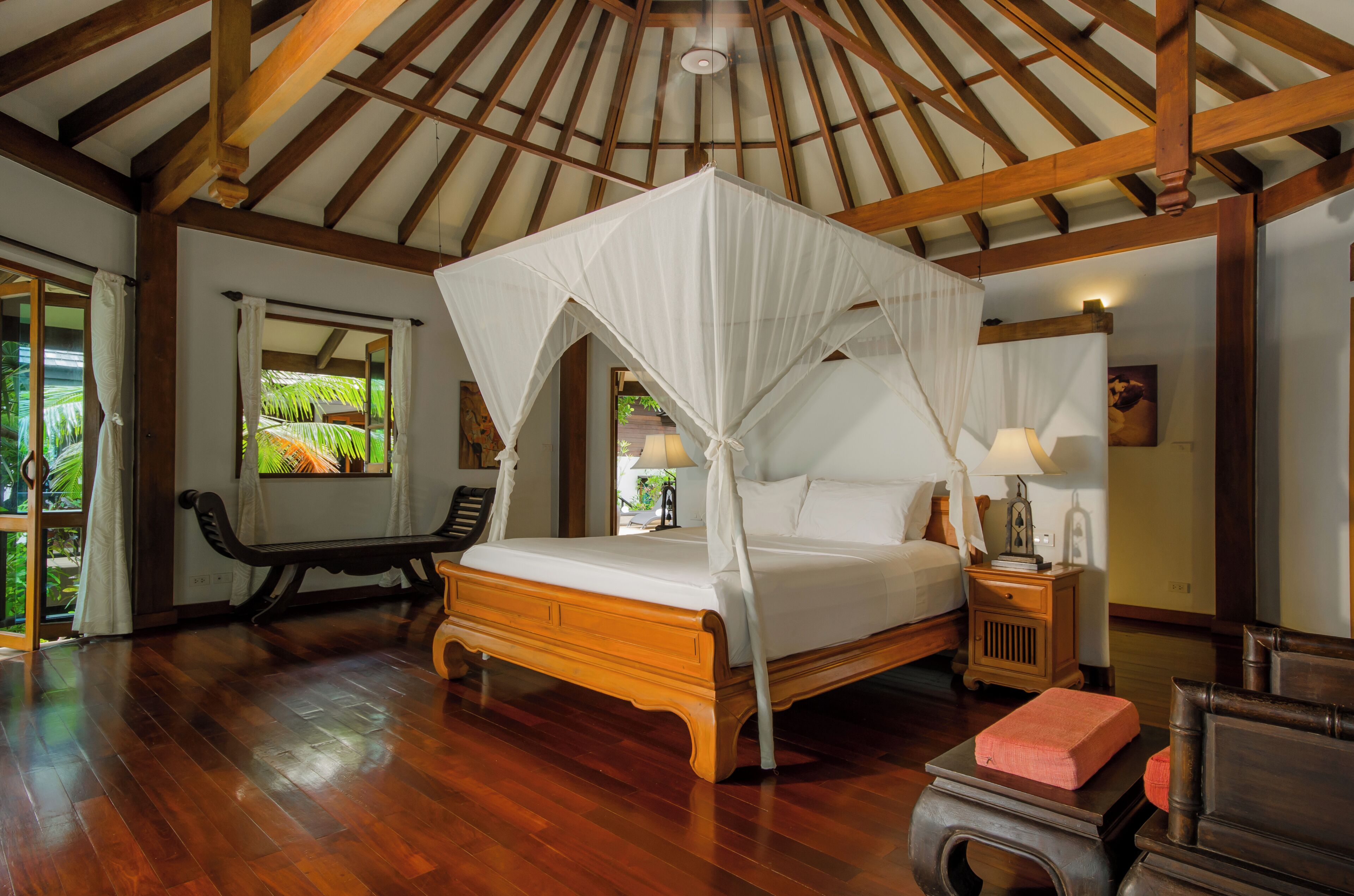 A bedroom with a wooden bed, a canopy bed, a bench, a nightstand, and a view of greenery outside.