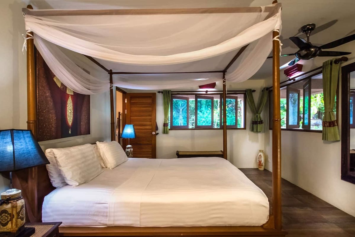 Un lit à baldaquin, une table de chevet avec une lampe, une tête de lit en bois, une fenêtre avec des rideaux et un ventilateur de plafond.