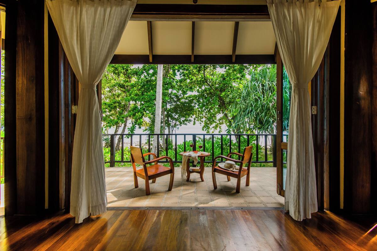 Un espace extérieur abrité avec des chaises et une table en bois, une vue sur la verdure à travers une rambarde et un plancher en bois.