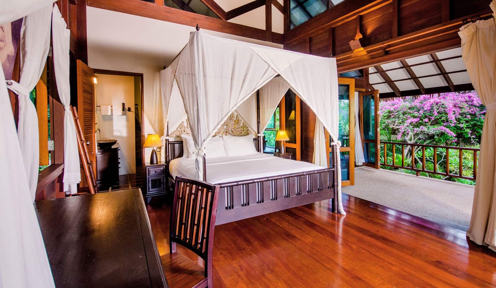 A room with a four-poster bed, wooden flooring, a nightstand, a lamp, and a view of greenery through a balcony.