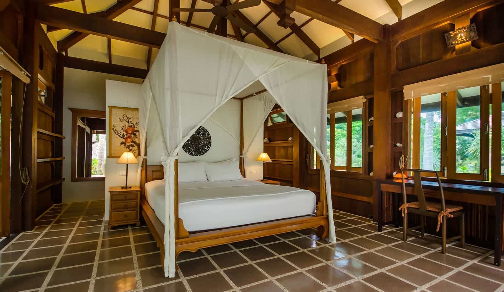 A four-poster bed with a canopy, wooden flooring, a desk with chairs, and a window with a view.