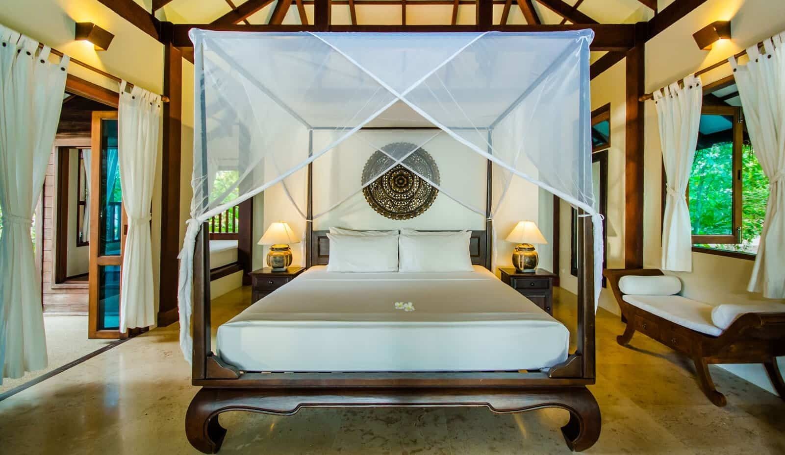 A four-poster bed with a canopy, two bedside lamps, a decorative wall hanging, and a wooden bench.