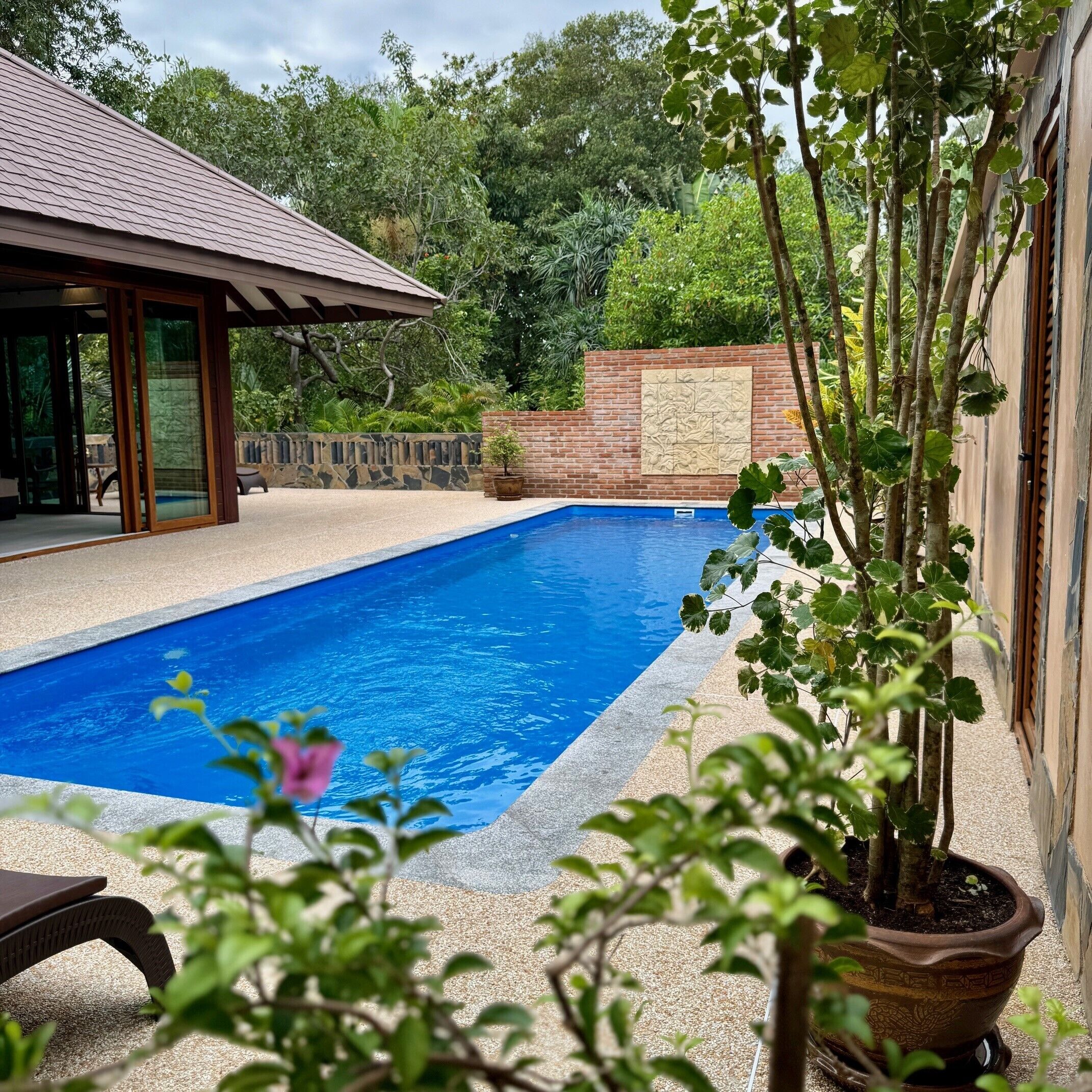 A swimming pool with a tiled deck, surrounded by a brick wall and lush greenery.