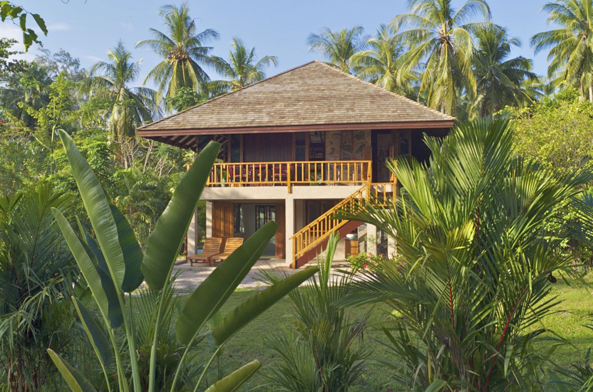 Casa de madera de dos pisos con balcón, rodeada de vegetación tropical.