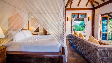 A bedroom with a canopy bed, a sofa with cushions, a window with a view of greenery, and a wooden ceiling structure.