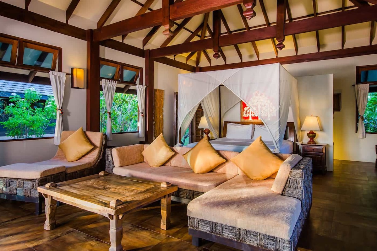 Une pièce avec un plafond en bois, un lit à baldaquin, un ensemble de canapés, une table basse en bois et des fenêtres avec des rideaux.