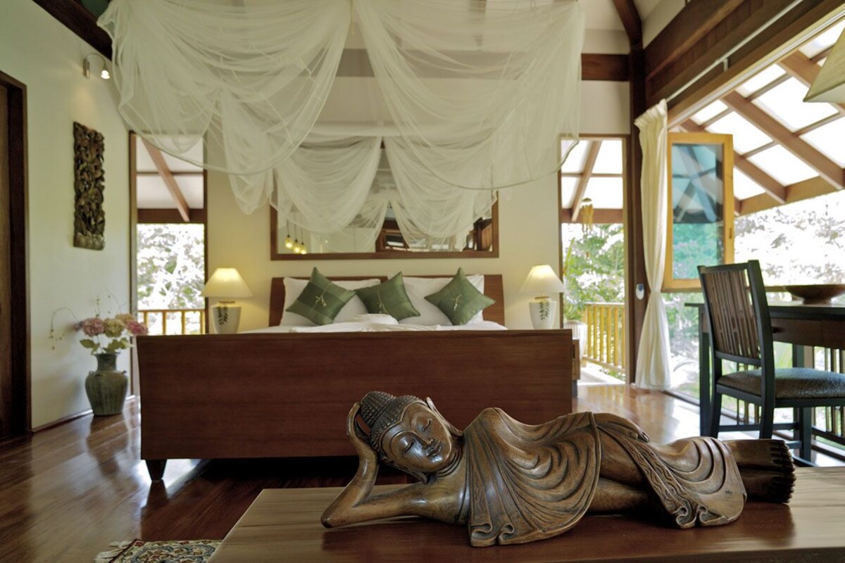 Une chambre à coucher comprenant un lit, une table de chevet, une chaise, une statue de Bouddha décorative et une fenêtre donnant sur un beau paysage.