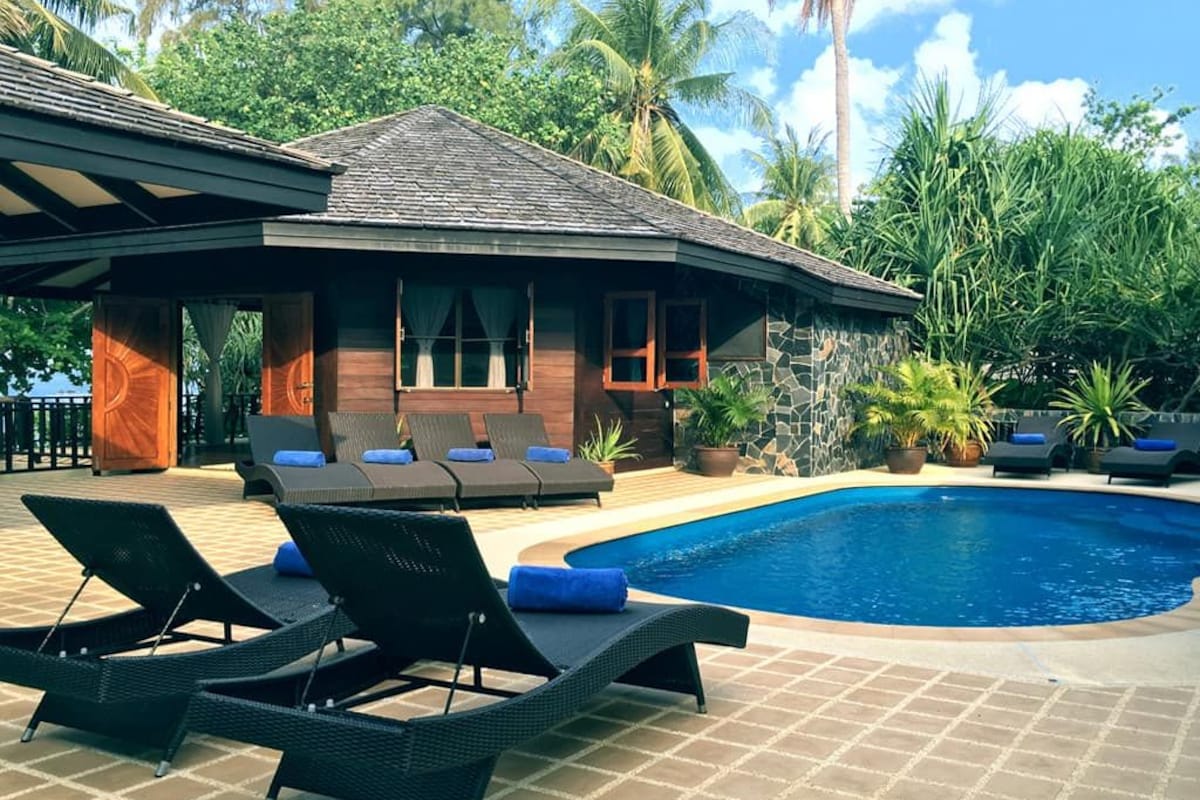 Un espace aménagé au bord de la piscine, avec des chaises longues, un bâtiment en pierre et une piscine d’eau cristalline, le tout entouré d’une végétation tropicale.