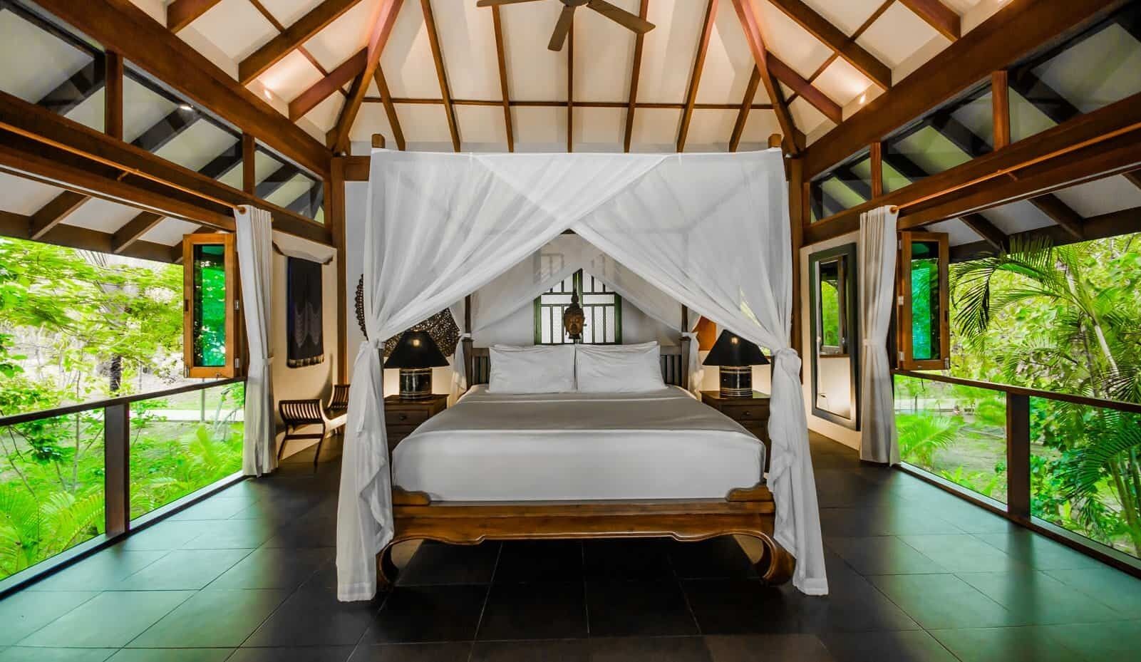 A bed with a canopy, a nightstand, a lamp, and a view of greenery through large windows.