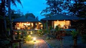 Un bâtiment à deux étages, doté d’un toit de chaume et entouré d’une végétation tropicale.