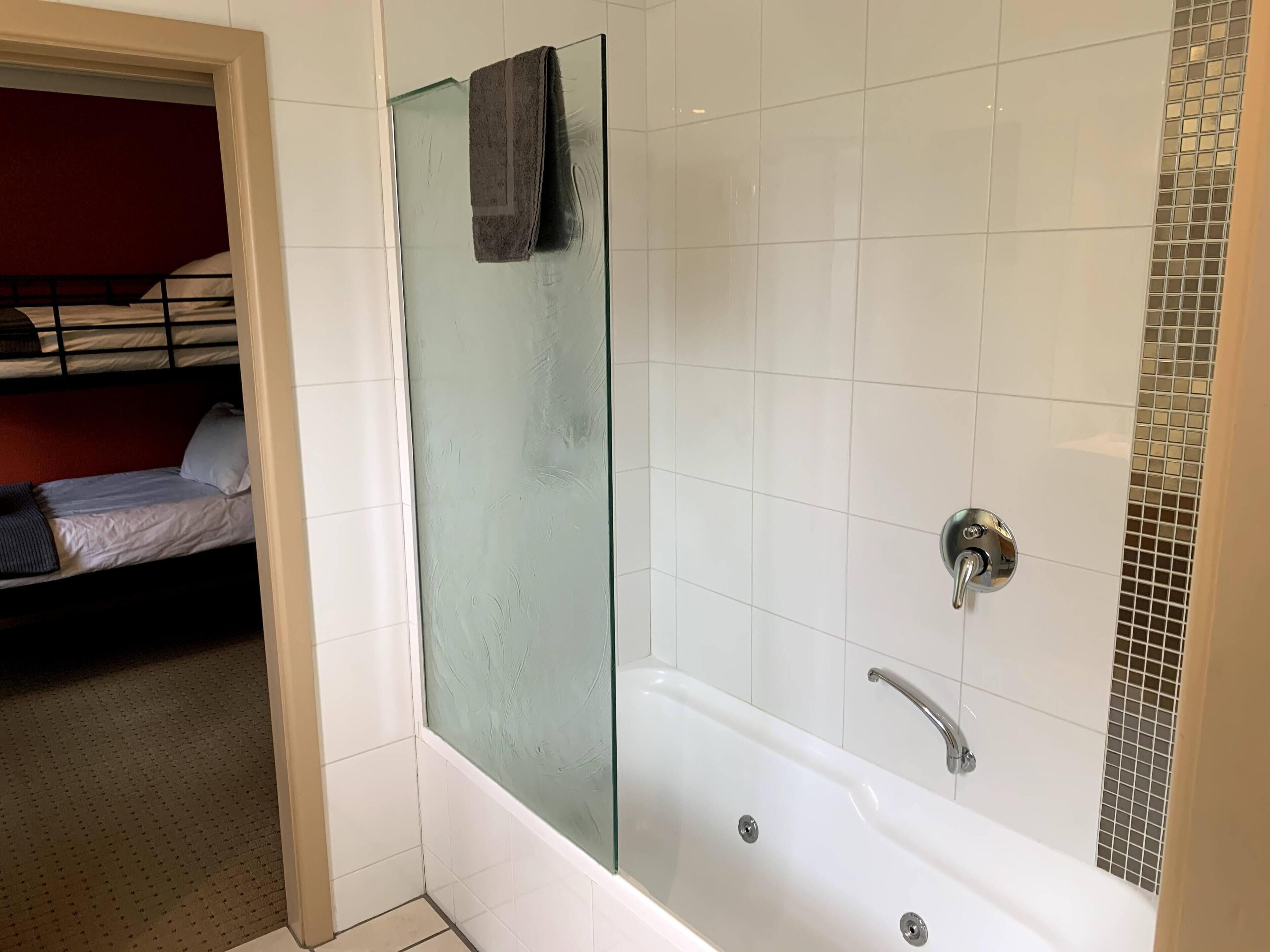 Une salle de bain avec une douche à parois de verre, une baignoire équipée d’un pommeau de douche et un lit dans la pièce adjacente.
