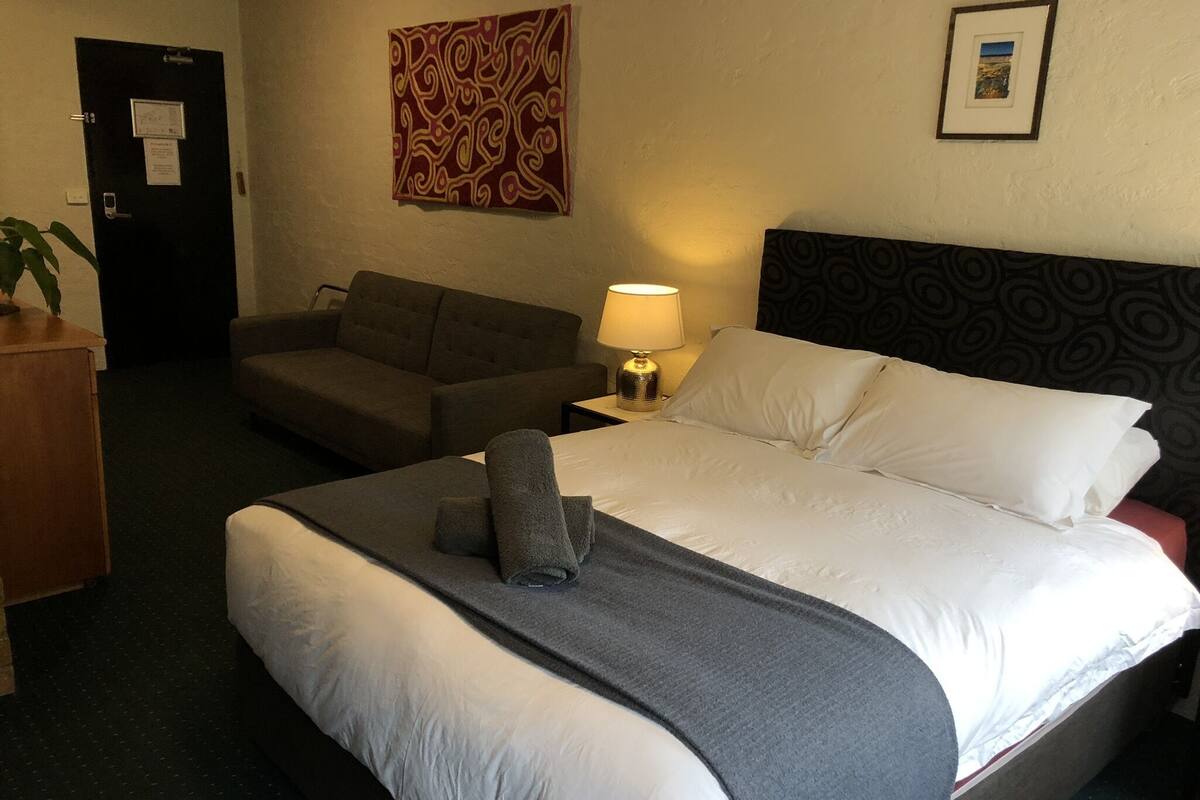 A hotel room with a large bed, a sofa, a desk with a lamp, and a door.
