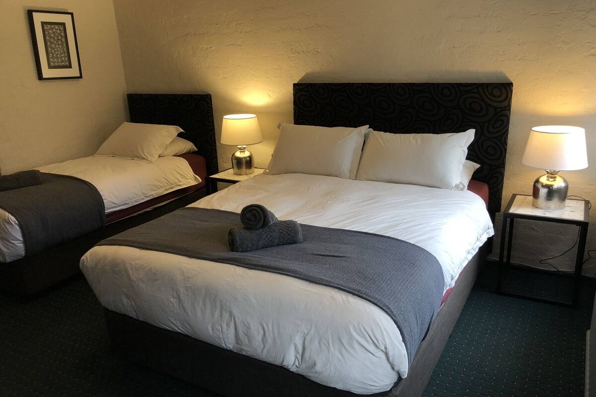 A hotel room with two beds, each with white linens and a dark headboard. There are two bedside tables with lamps, and a framed picture on the wall.