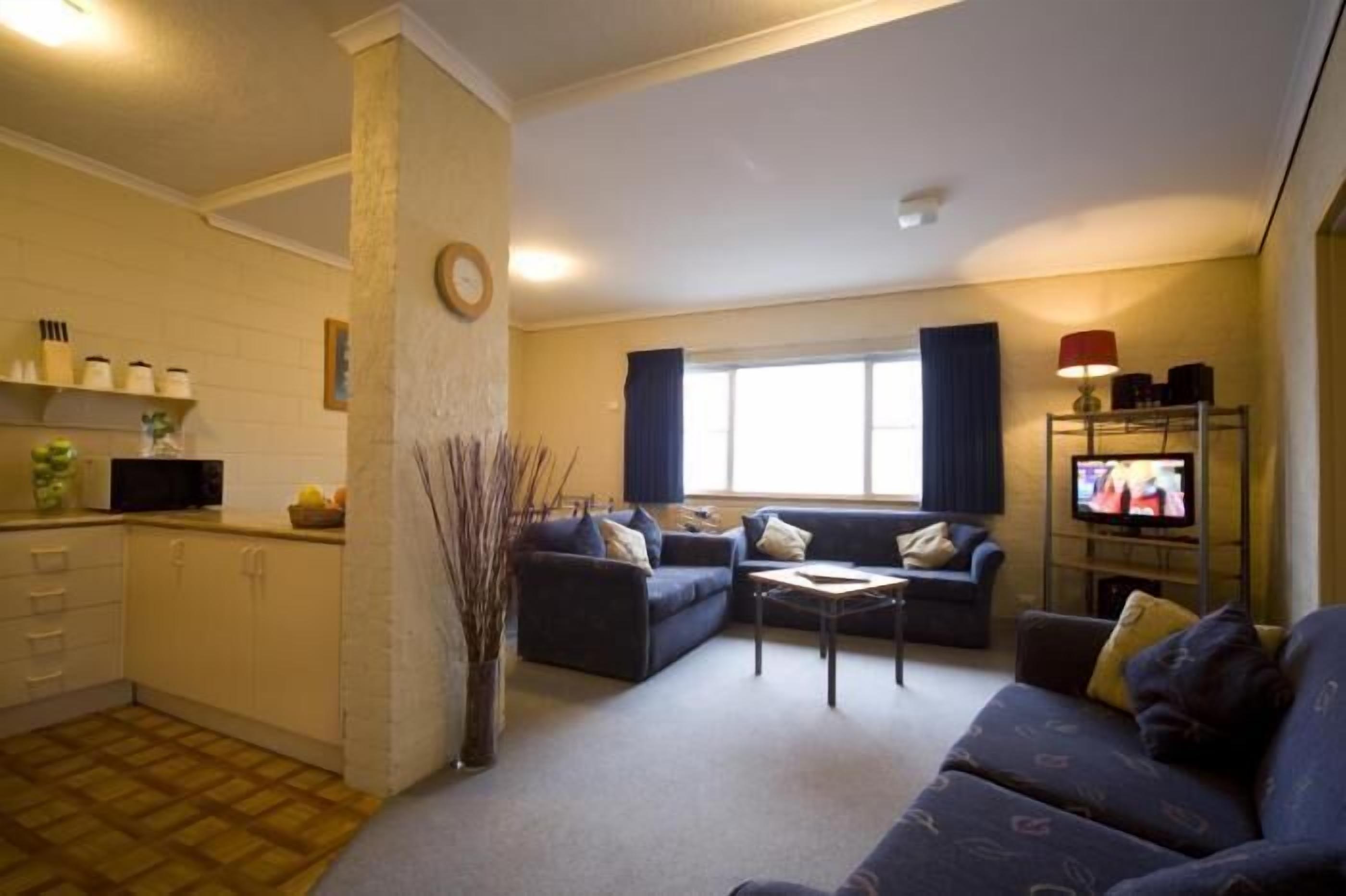 A living room with a sofa, a coffee table, and a TV. There is a kitchen area with cabinets and a microwave.