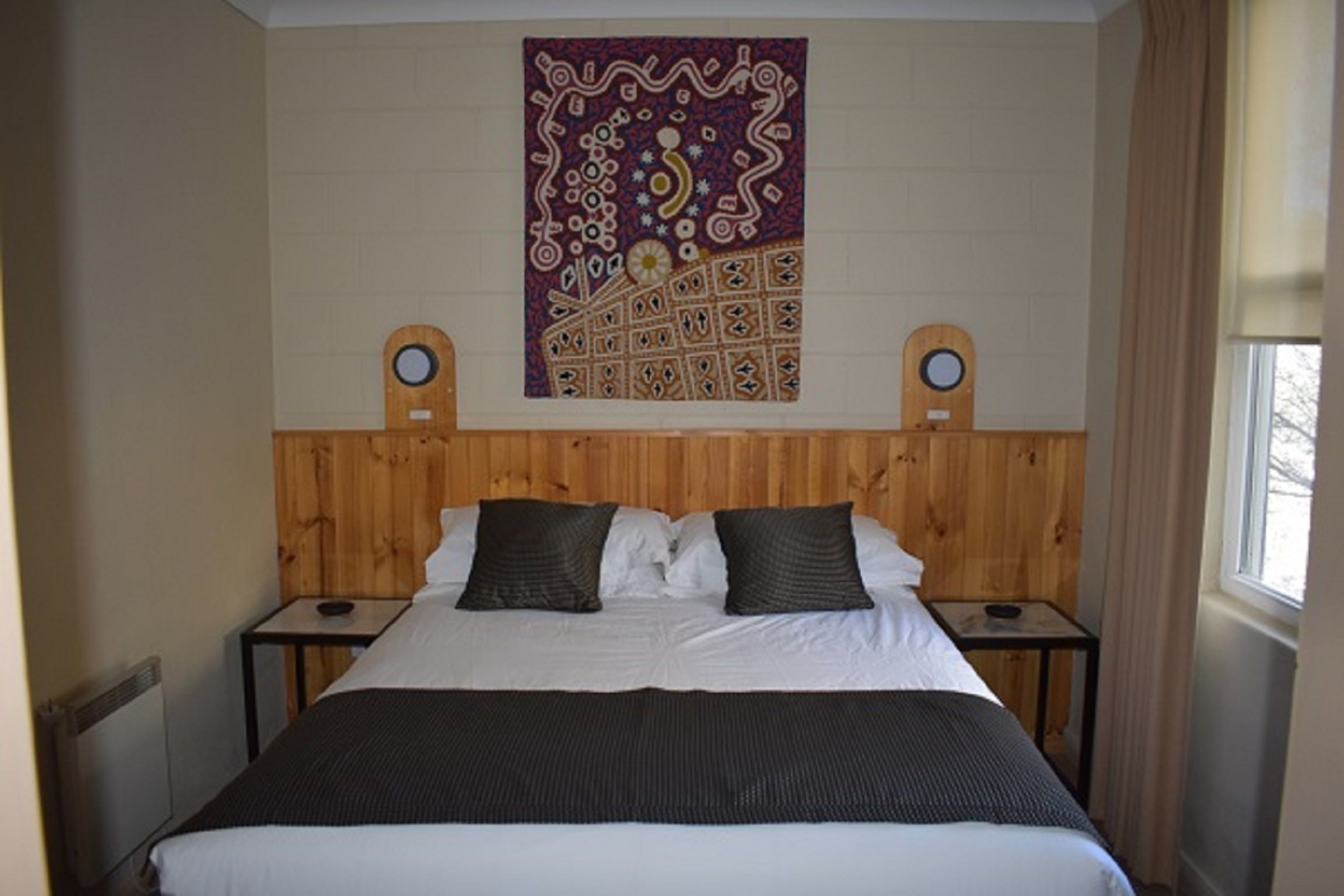 A bedroom with a wooden headboard, two nightstands, a bed with white linens and two dark pillows, and a tapestry on the wall.