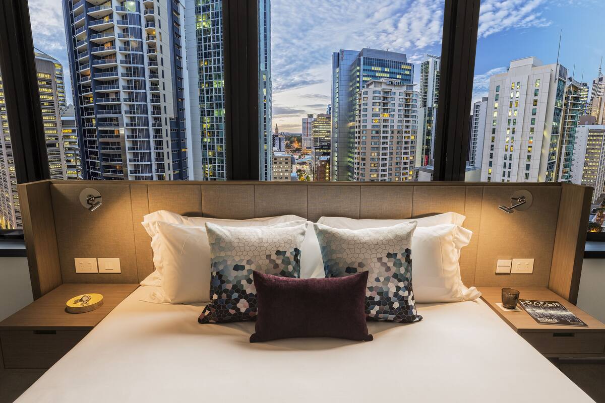 Habitación de hotel con una cama grande, dos mesitas de noche y vistas de la ciudad a través de amplios ventanales.