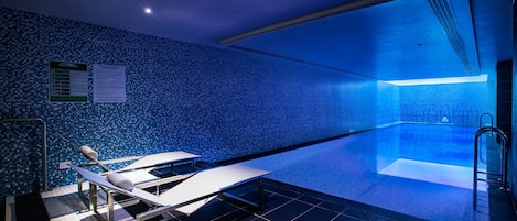 Una piscina interior moderna con pared de azulejos azules, un mostrador de recepción y una zona iluminada por el sol.