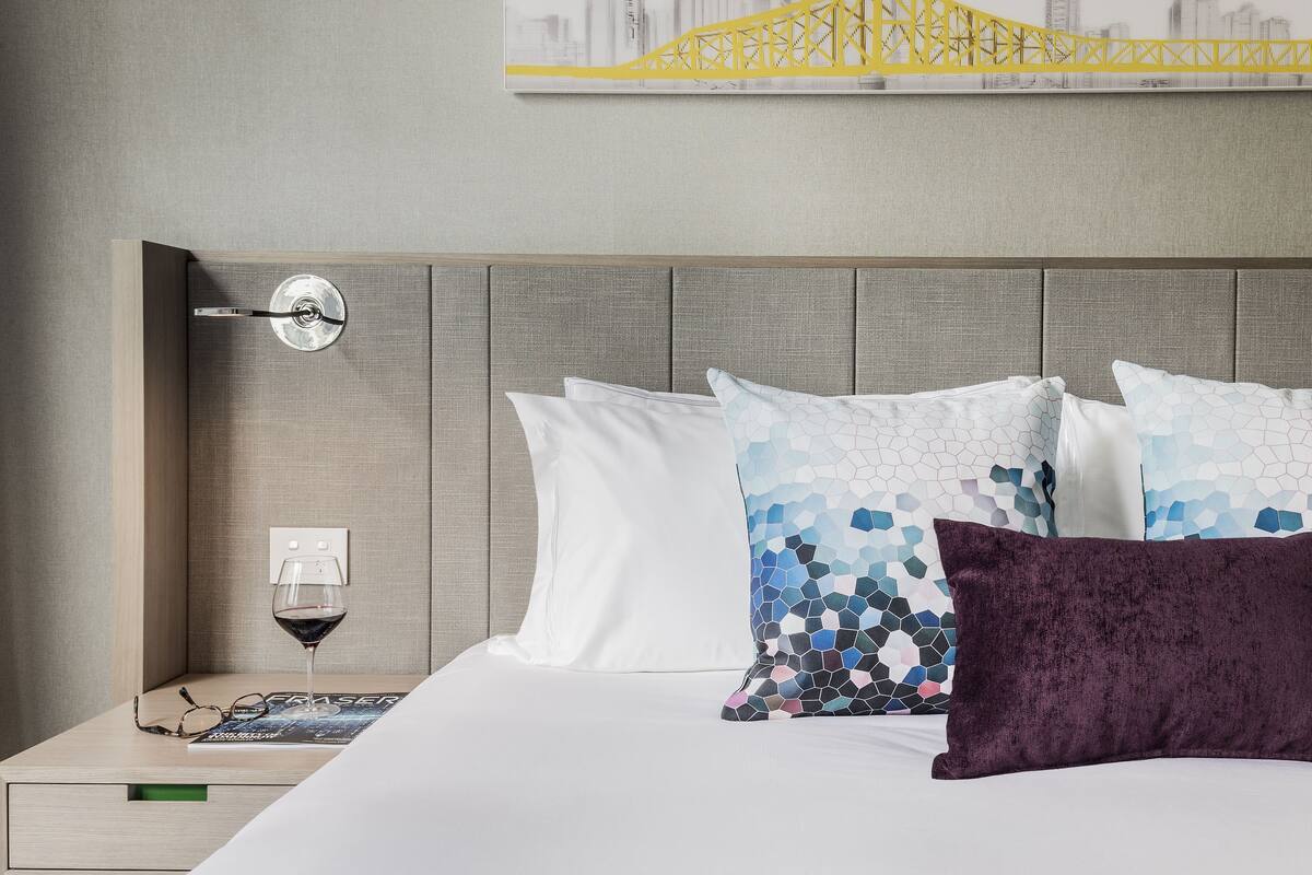 Una habitación de hotel con una cama, una mesita de noche, una copa de vino y una obra de arte con un paisaje urbano en la pared.