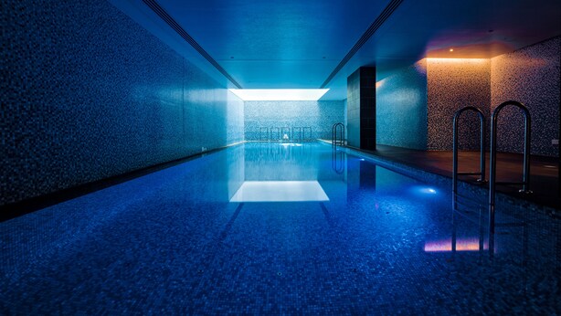 A modern indoor swimming pool with a blue mosaic floor and walls, a central light fixture, and metal railings along the sides. - Capri by Fraser, Brisbane (Brisbane)