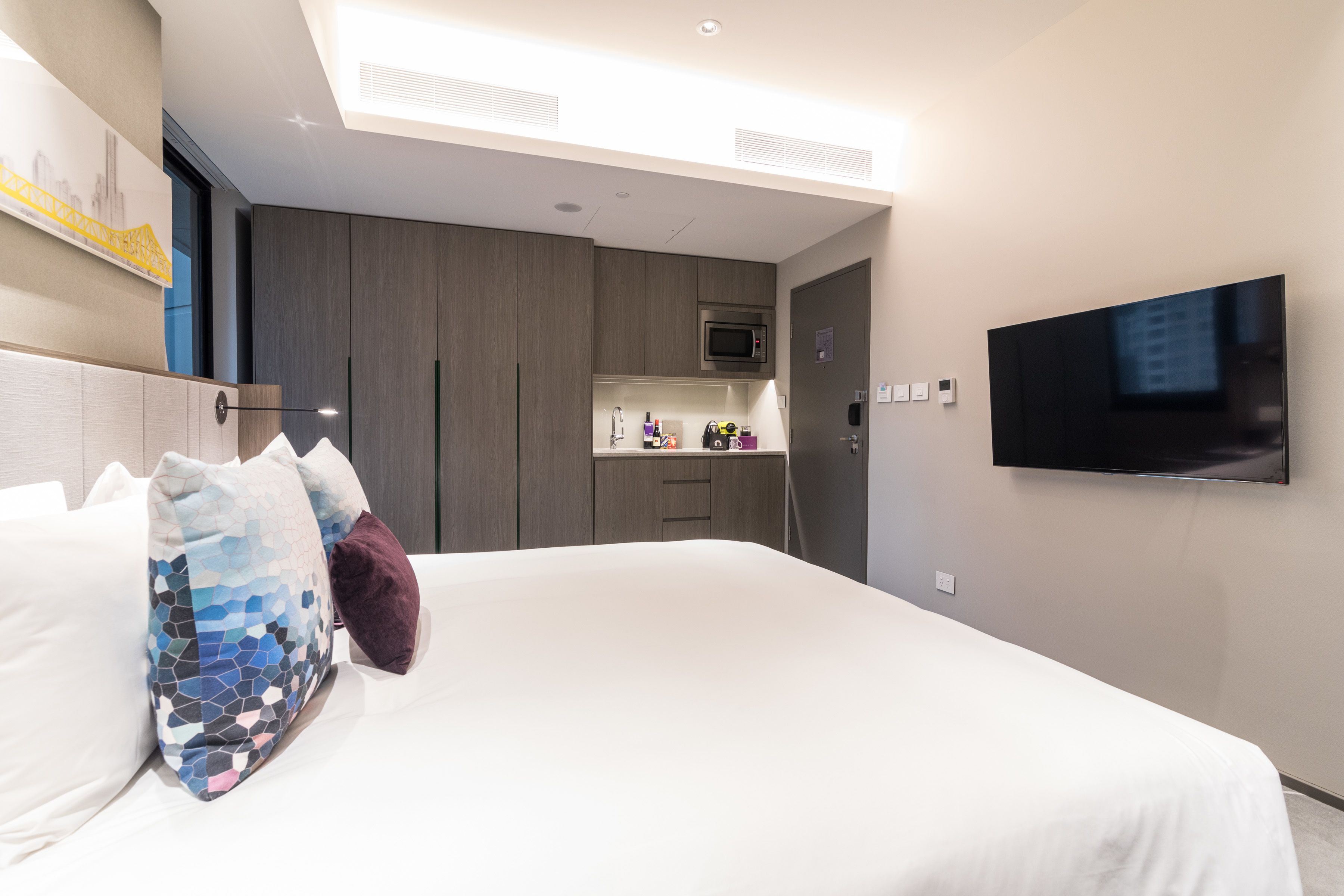 Una habitación de hotel moderna con una cama grande, un televisor de pantalla plana montado en la pared y una kitchenette con microondas y refrigerador pequeño.