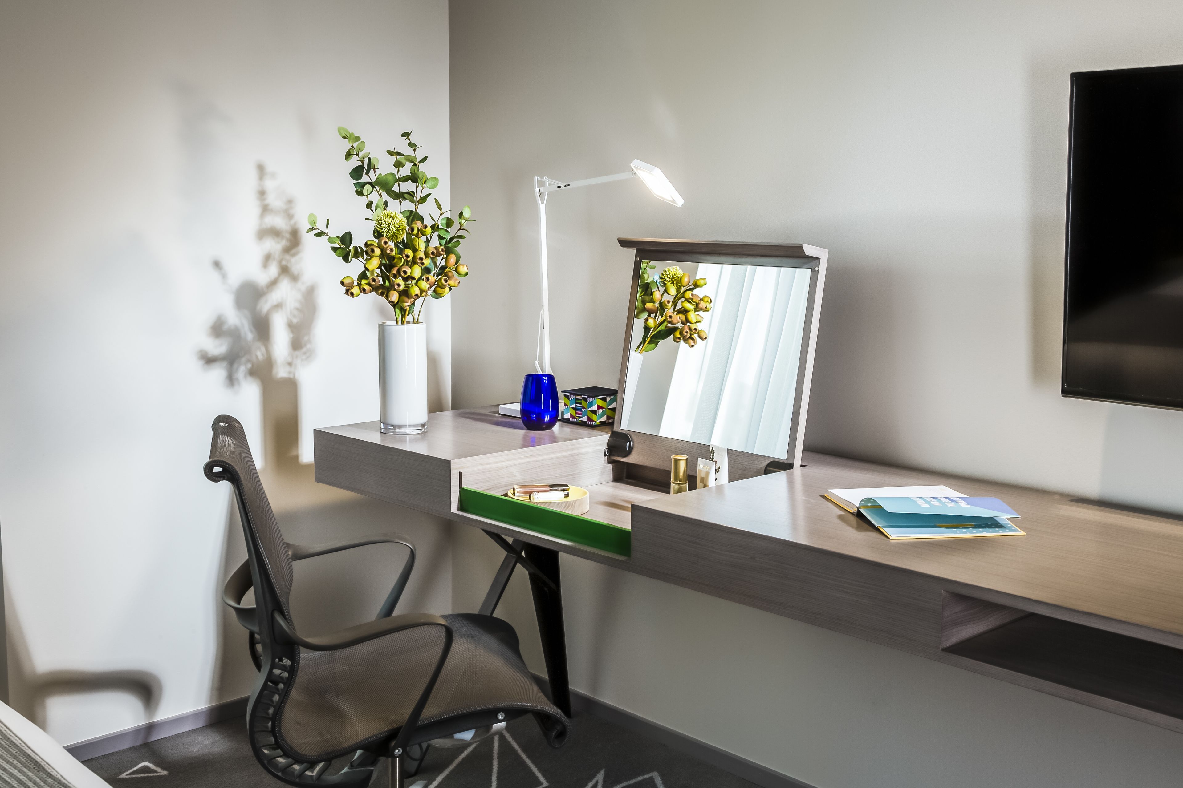 A modern office desk with a chair, a desk lamp, a vase with flowers, a mirror, and a flat-screen TV.
