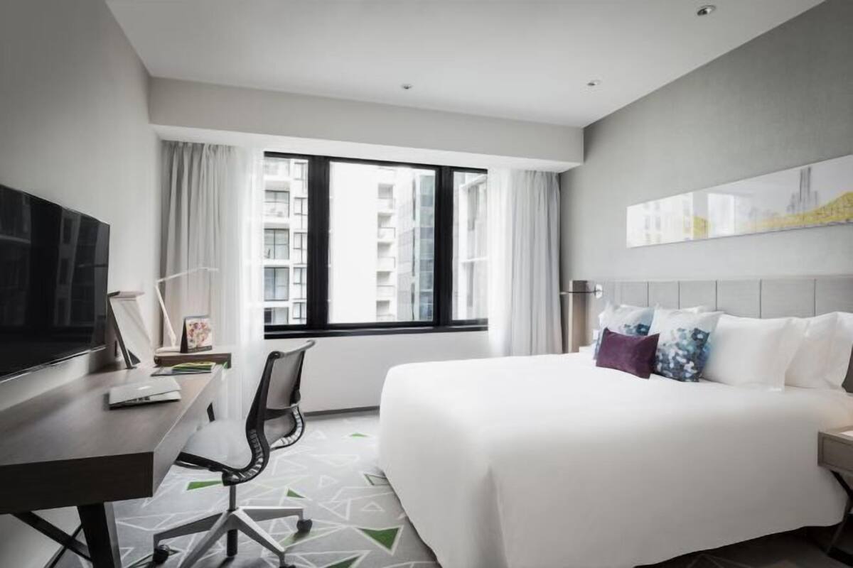 Habitación de hotel con cama, escritorio, silla, televisión y ventana con vista a edificios.