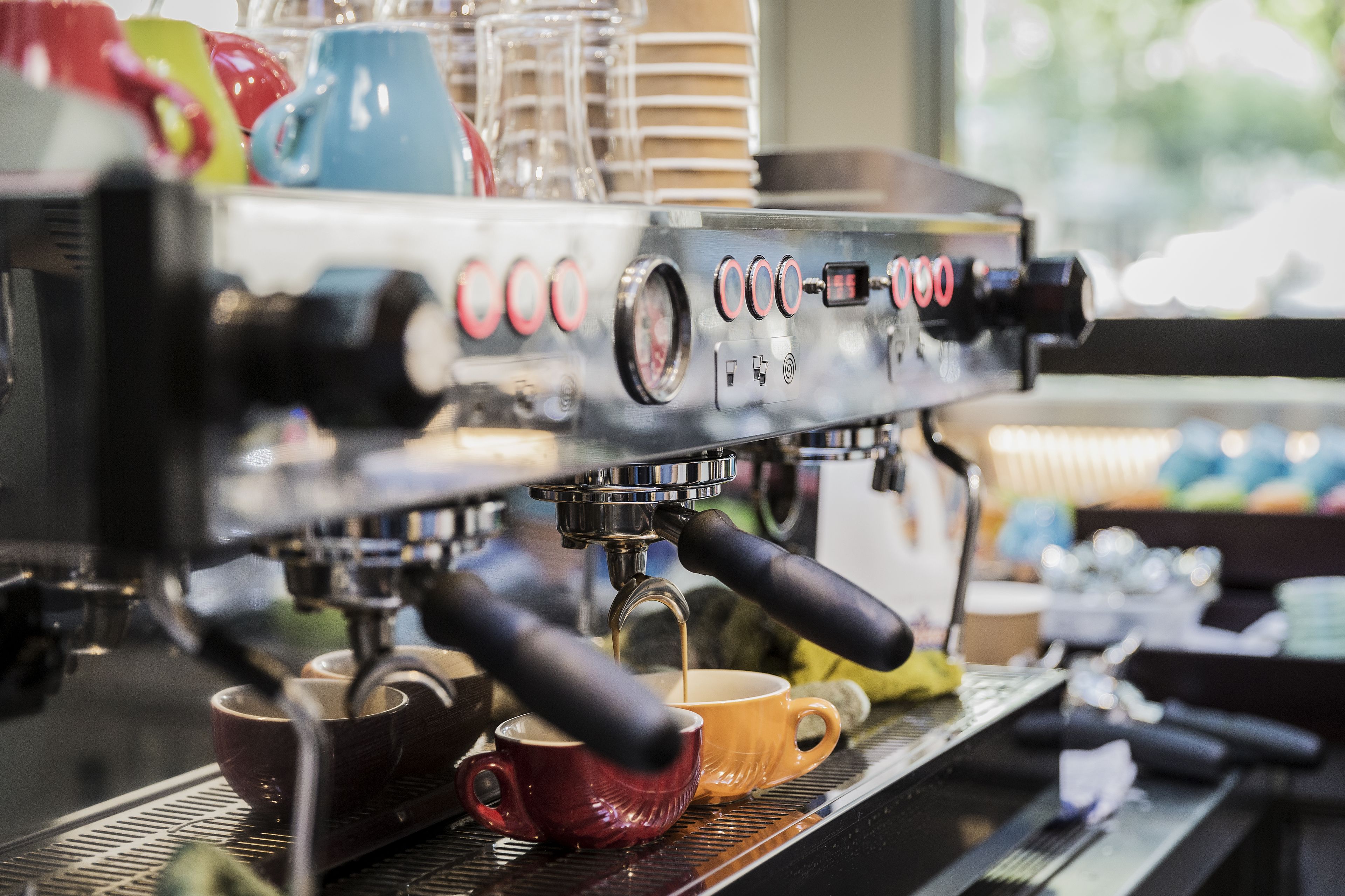 A coffee machine with multiple buttons and a digital display, cups placed below it, and a window in the background.