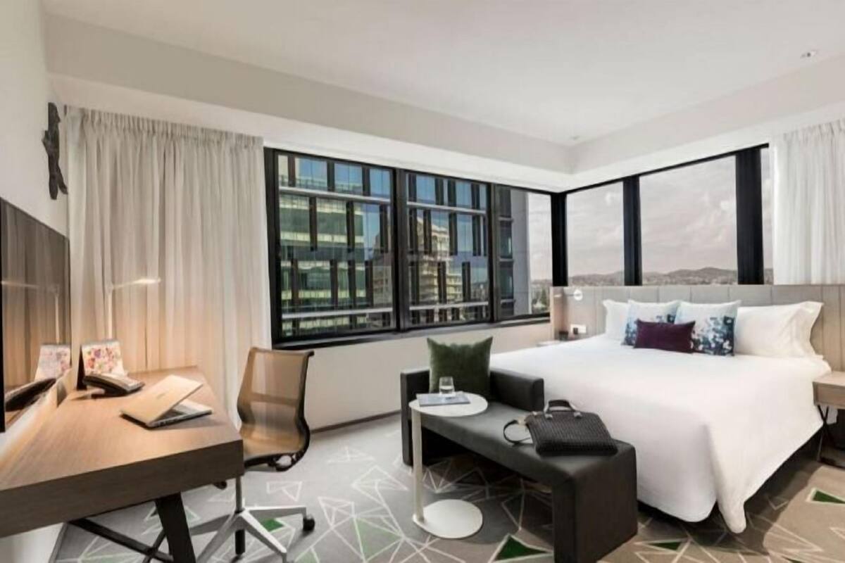 Una habitación de hotel moderna con una cama grande, un escritorio, una silla y vistas de la ciudad a través de amplios ventanales.