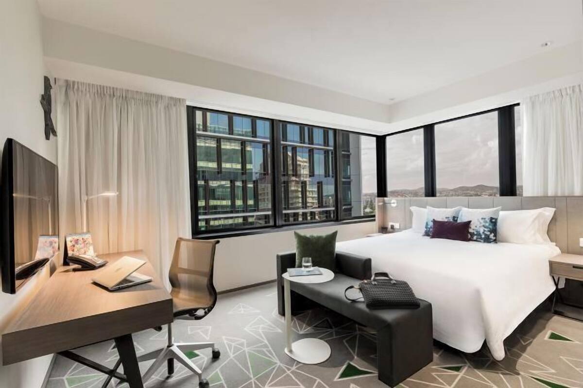 Un hotel moderno con una cama grande, un escritorio con una computadora portátil, una silla y vistas de la ciudad a través de amplias ventanas.