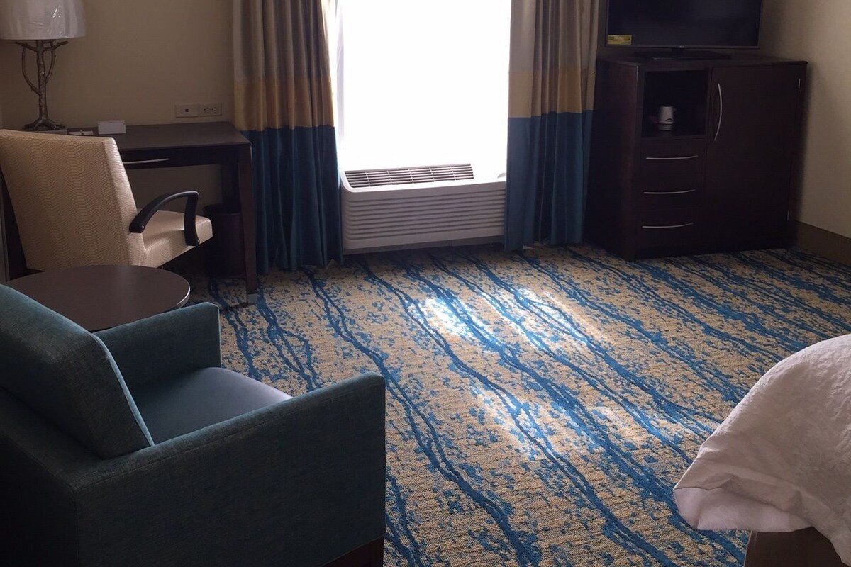 A hotel room with a desk, chair, TV, and a large window with curtains.