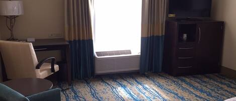 A hotel room with a desk, chair, TV, and a large window with curtains.