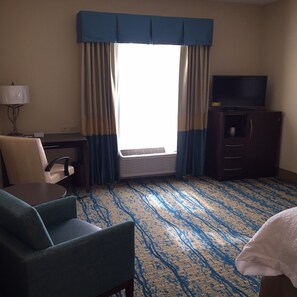 Habitación de hotel con escritorio, silla, televisor y una ventana grande con cortinas.