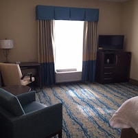 A hotel room with a desk, chair, TV, and a large window with curtains.