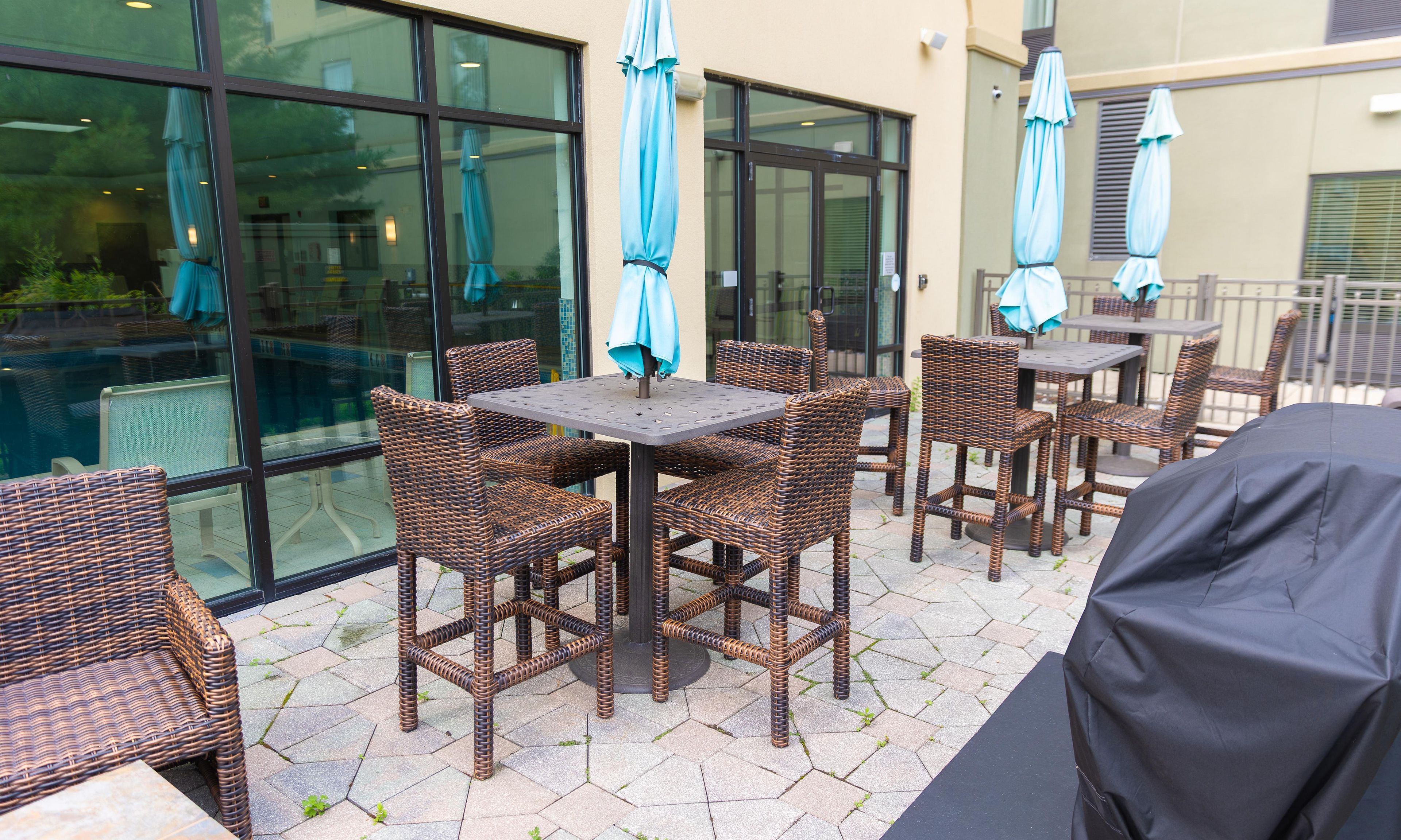 An outdoor patio area with wicker furniture and umbrellas, a covered grill, and a view of the building's interior through glass doors.