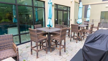 Een terras buiten met rieten meubilair en parasols, een overdekte barbecue en uitzicht op het interieur van het gebouw door glazen deuren.